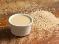 Un cuenco blanco con cremosa salsa de tahini junto a una pila de semillas de sésamo sobre una tabla de madera.