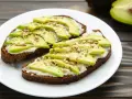 Dos tostadas de pan integral con queso crema, rodajas de aguacate y semillas. Un desayuno saludable y delicioso.