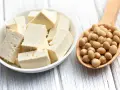 Tofu en cubos en un plato blanco junto a una cuchara de madera llena de semillas de soja. ¿A que sabe el tofu? Neutro, absorbe sabores.