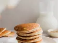 Una pila de esponjosos **pancakes de avena** dorados, con un jarro de leche y un tazón de canela al fondo.