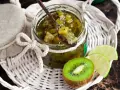 Tarro de mermelada de kiwi casera, con una cuchara de madera llena de la dulce conserva. Rodajas de kiwi y lima acompañan la presentación.