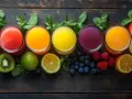Cinco vasos de zumos de colores vibrantes, rodeados de frutas frescas como limones, kiwis, fresas, frambuesas y arándanos. Una opción saludable de zumos para la anemia.