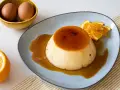 Delicioso flan de naranja con caramelo líquido, decorado con rodajas y ralladura de naranja. Huevos en un cuenco al fondo.