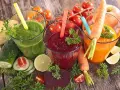 Tres zumos astringentes de colores vibrantes: verde, rojo y naranja, rodeados de ingredientes frescos como zanahorias, remolachas y tomates.