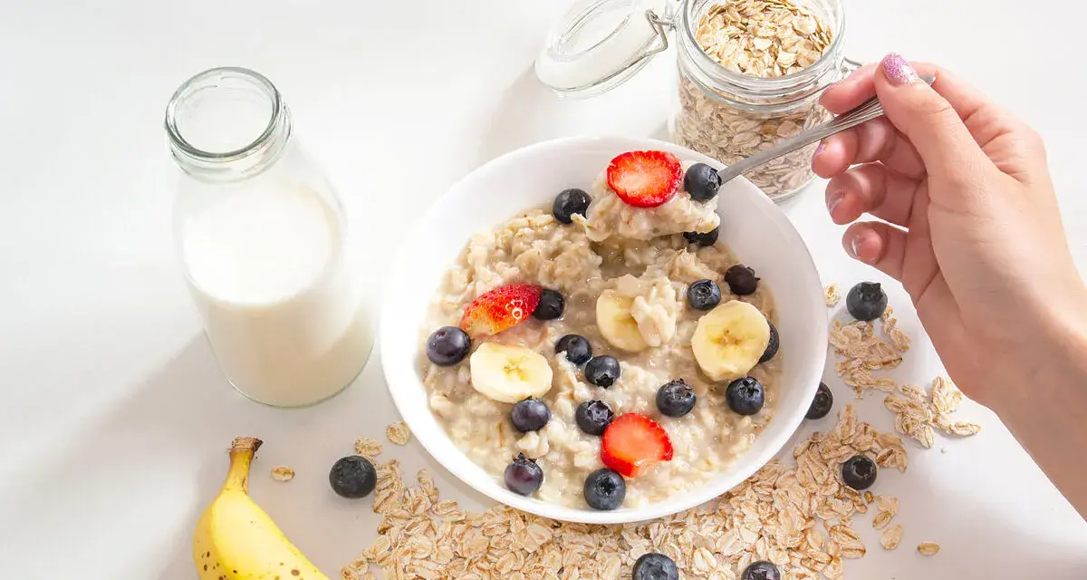 Un tazón de avena con fresas, arándanos y plátano. Un frasco de avena y una botella de leche sugieren cómo se hacen los copos de avena.