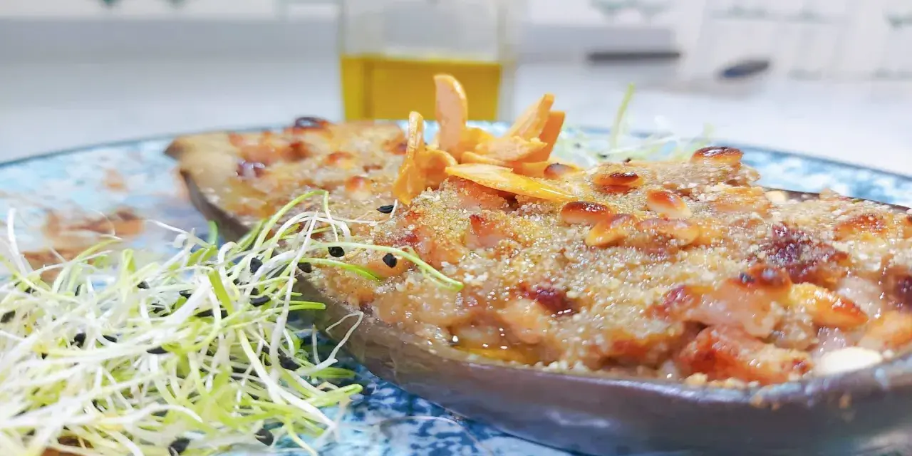 Deliciosa berenjena rellena gratinada, adornada con piñones y brotes frescos, lista para ser disfrutada.