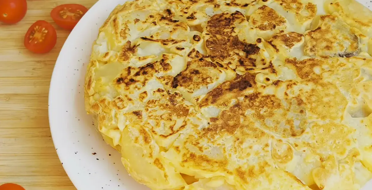Tortilla vegana dorada y esponjosa, con trozos de patata visibles, servida en plato blanco. Tomates cherry al lado.