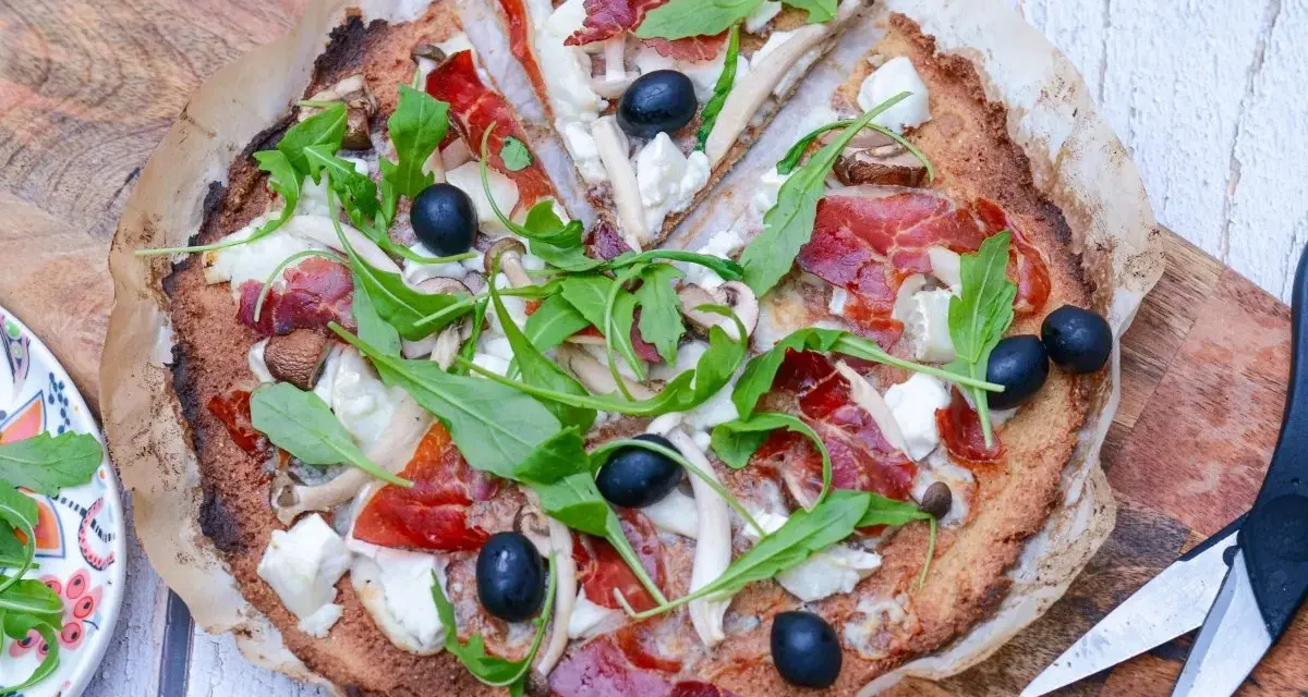 Pizza de quinoa con jamón serrano, aceitunas negras, champiñones y rúcula fresca. Una delicia saludable.