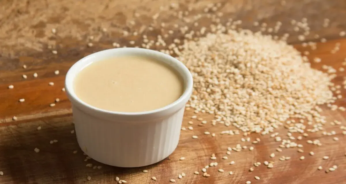 Un cuenco blanco con cremosa salsa de tahini junto a una pila de semillas de sésamo sobre una tabla de madera.