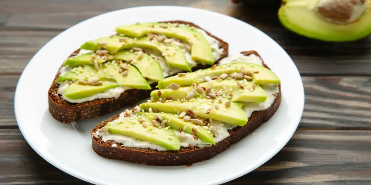 Dos tostadas de pan integral con queso crema, rodajas de aguacate y semillas. Un desayuno saludable y delicioso.