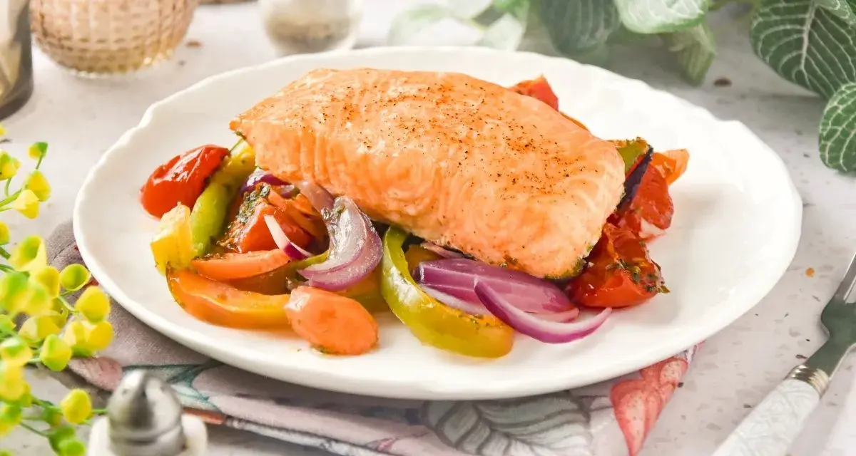 Un jugoso salmón al horno reposa sobre una cama de verduras asadas: tomates, pimientos y cebolla morada.
