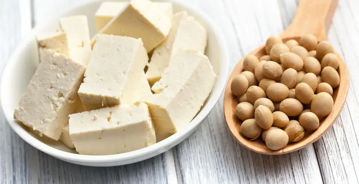 Tofu en cubos en un plato blanco junto a una cuchara de madera llena de semillas de soja. ¿A que sabe el tofu? Neutro, absorbe sabores.