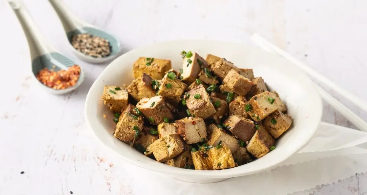Tazón lleno de cubos dorados de tofu marinado, espolvoreados con cebollín y semillas.