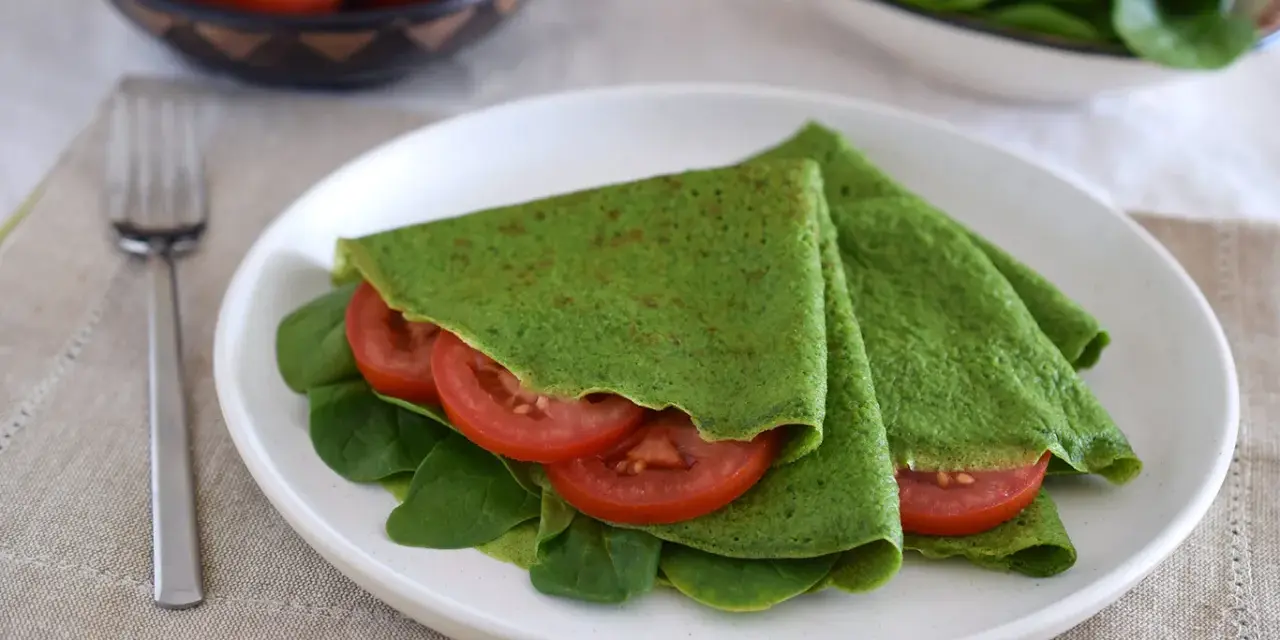 Deliciosos crepes de espinacas rellenos de rodajas de tomate fresco y hojas de espinaca, servidos en un plato blanco.