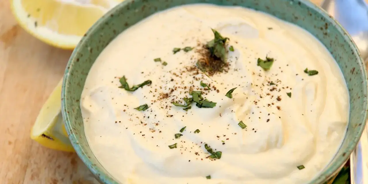 Un cremoso tazón de mayonesa de tofu, adornado con perejil fresco y pimienta negra, junto a rodajas de limón.