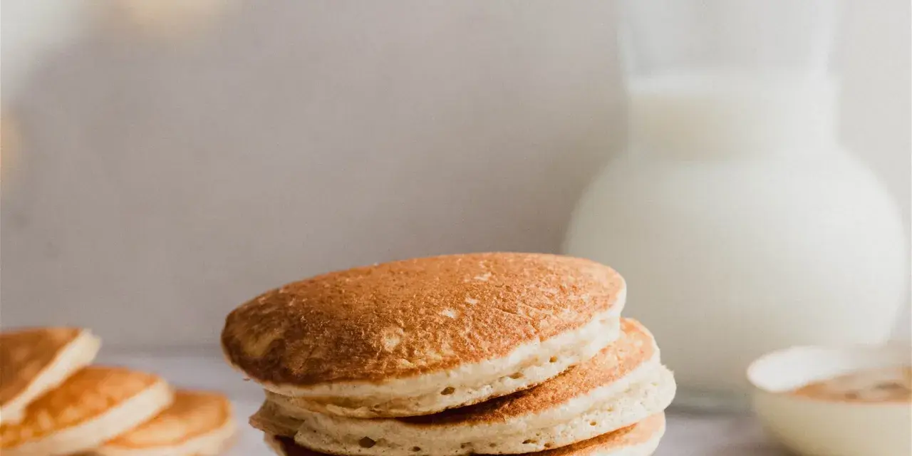 Una pila de esponjosos **pancakes de avena** dorados, con un jarro de leche y un tazón de canela al fondo.