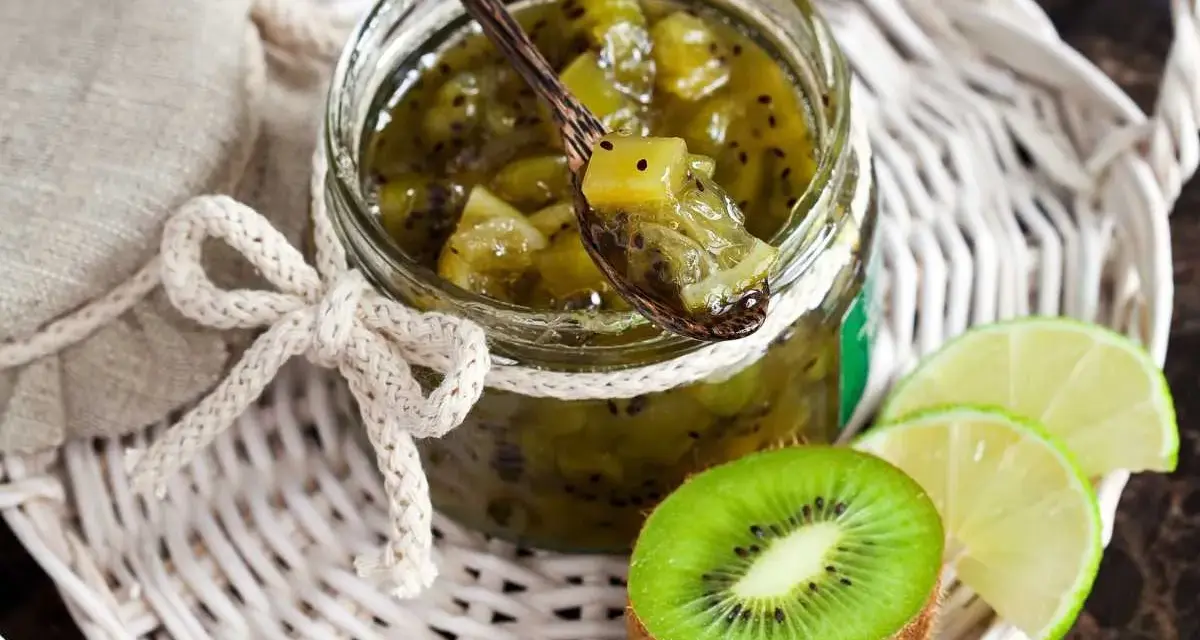Tarro de mermelada de kiwi casera, con una cuchara de madera llena de la dulce conserva. Rodajas de kiwi y lima acompañan la presentación.