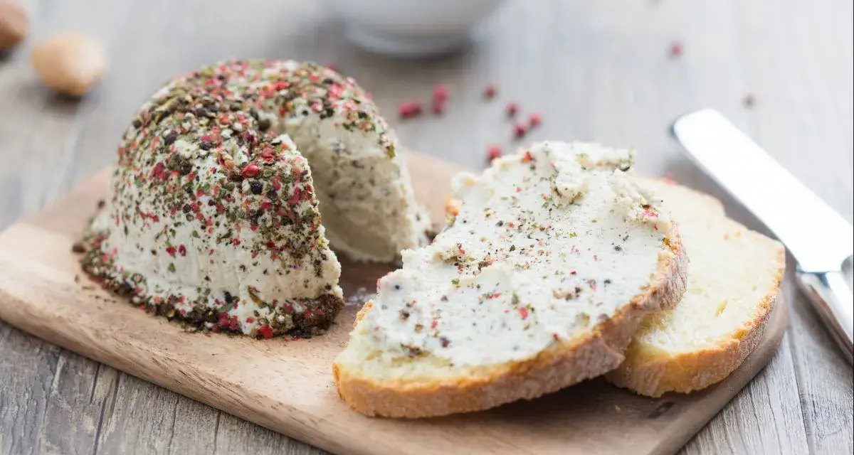 Queso vegano receta: un bloque de queso cremoso cubierto de pimienta y hierbas, servido con rebanadas de pan.