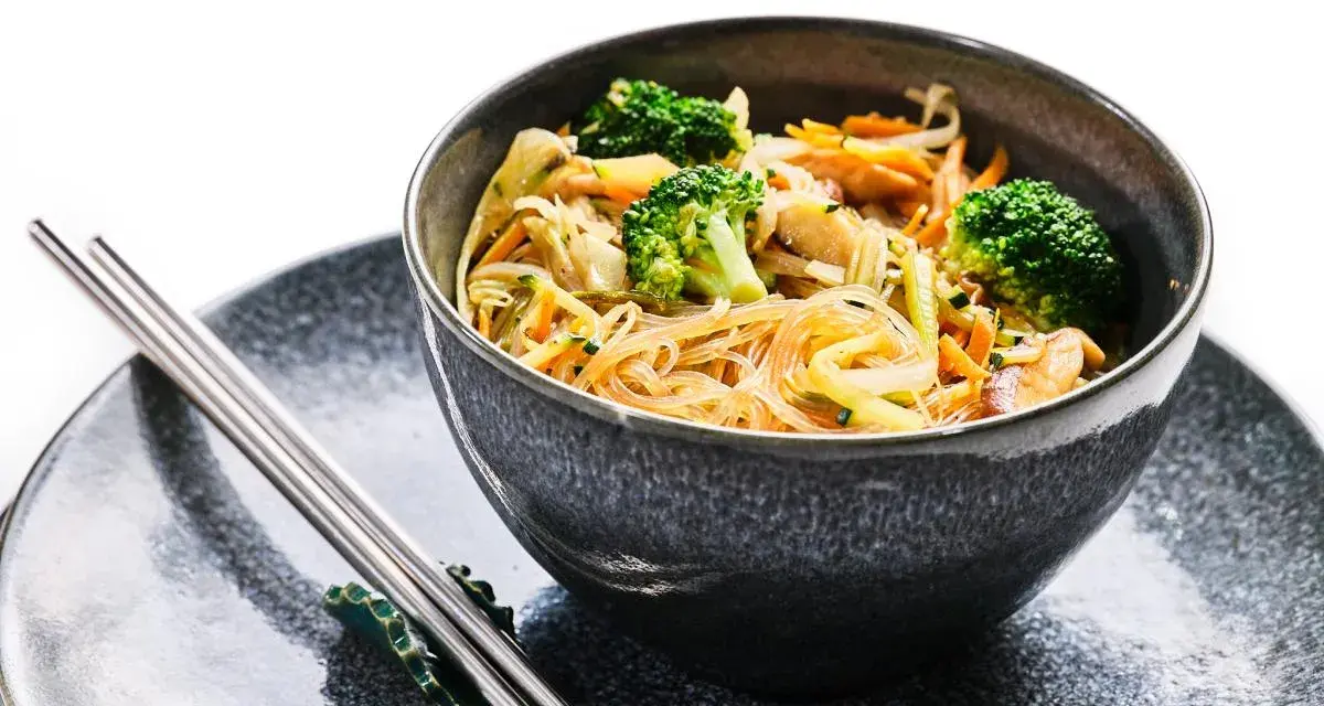 Deliciosa receta con noodles de arroz, brócoli, zanahoria y otros vegetales frescos en un tazón gris, acompañado de palillos.