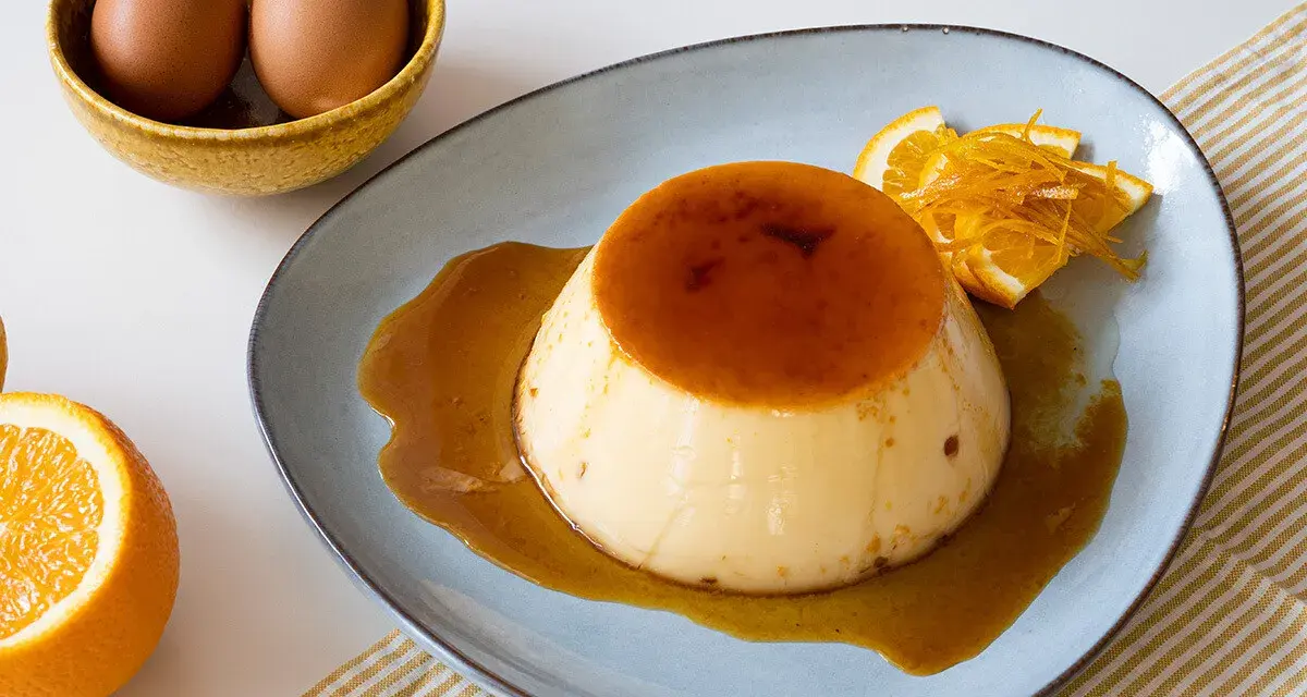 Delicioso flan de naranja con caramelo líquido, decorado con rodajas y ralladura de naranja. Huevos en un cuenco al fondo.