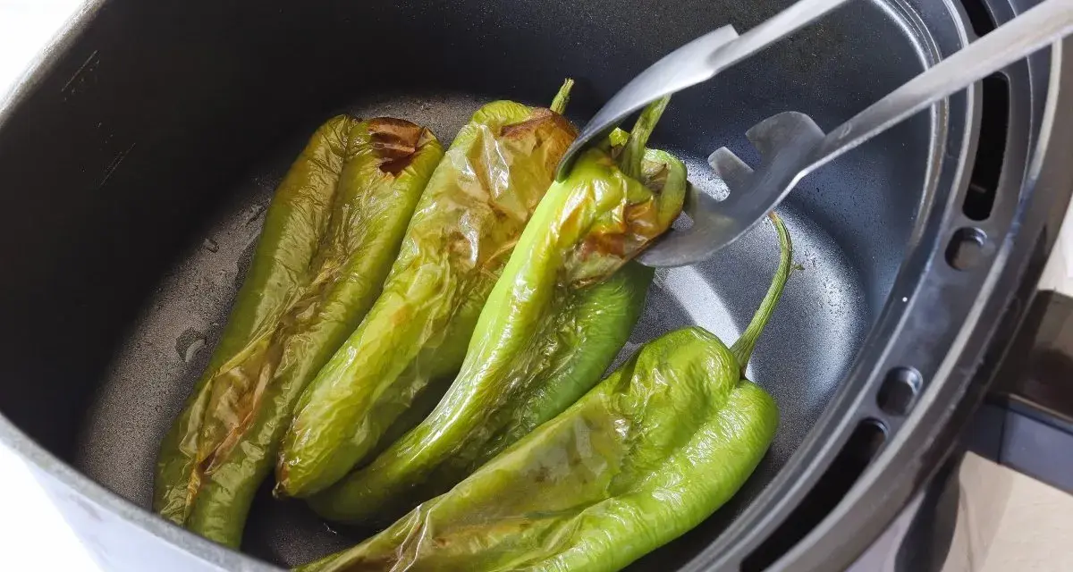 Pimientos verdes asándose en una freidora de aire. Unas pinzas metálicas sujetan uno de los pimientos, mostrando su piel ligeramente chamuscada.