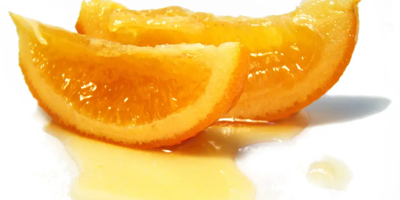 Dos rodajas de naranja confitada brillan con dulzura, bañadas en un almíbar dorado sobre fondo blanco.