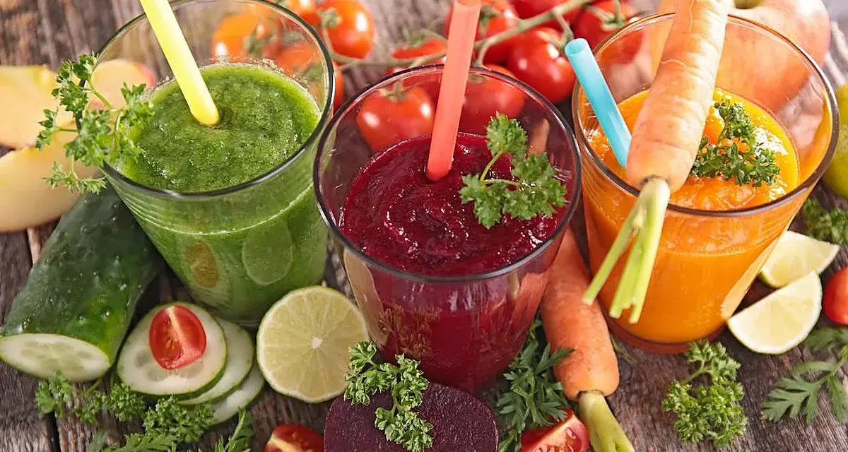 Tres zumos astringentes de colores vibrantes: verde, rojo y naranja, rodeados de ingredientes frescos como zanahorias, remolachas y tomates.