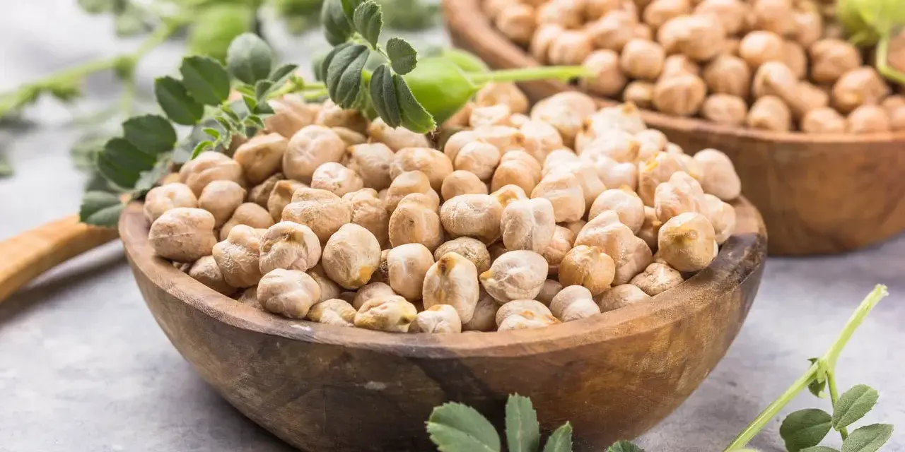 Dos cuencos de madera llenos de garbanzos secos, con hojas verdes y vainas. Los garbanzos tienen fibra, ¡un alimento saludable!