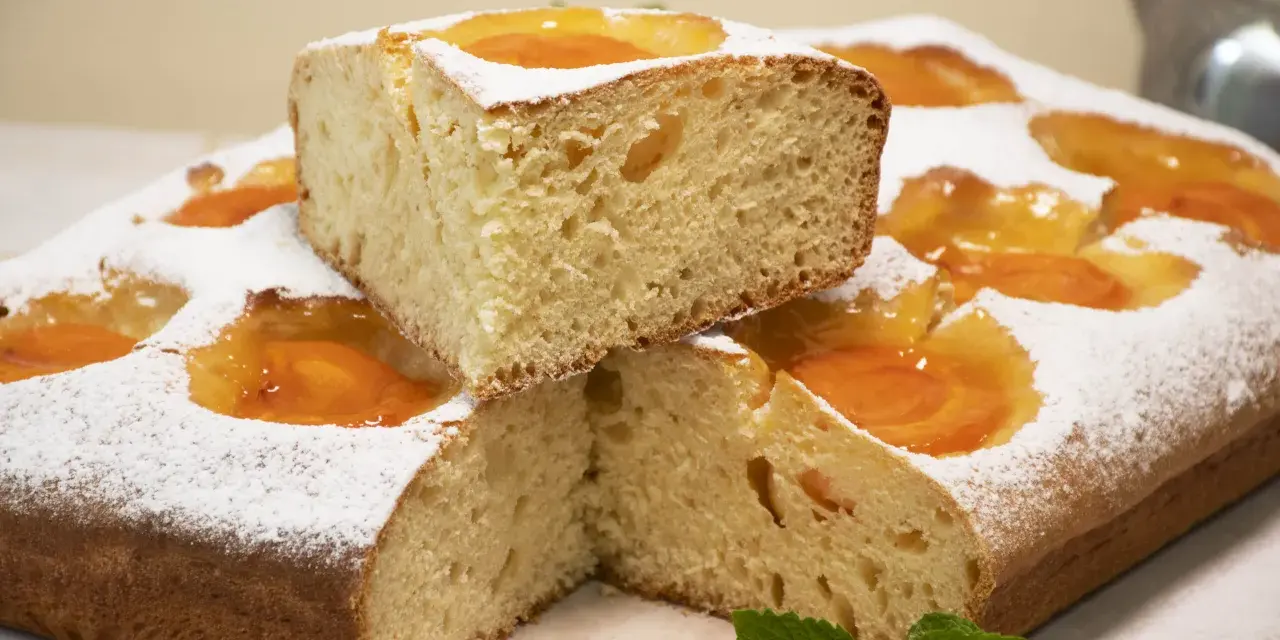 Deliciosa coca de albaricoque, cubierta de azúcar glas y decorada con trozos de fruta. Un trozo apilado revela su esponjosa miga.