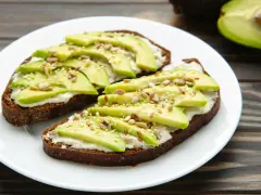 Dos tostadas de pan integral con queso crema, rodajas de aguacate y semillas. Un desayuno saludable y delicioso.