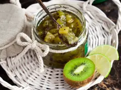 Tarro de mermelada de kiwi casera, con una cuchara de madera llena de la dulce conserva. Rodajas de kiwi y lima acompañan la presentación.