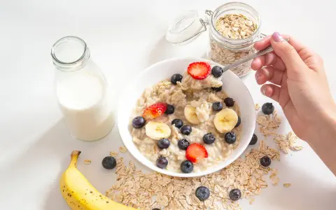 Un tazón de avena con fresas, arándanos y plátano. Un frasco de avena y una botella de leche sugieren cómo se hacen los copos de avena.