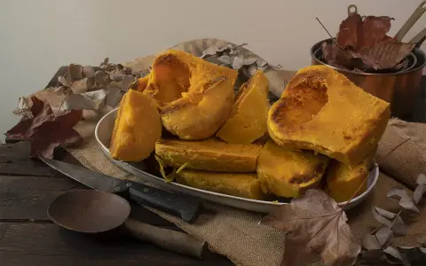 Trozos de calabaza asada, tierna y dorada, servidos en un plato blanco sobre arpillera. Acompañada de utensilios rústicos y hojas secas, evoca el sabor del otoño.