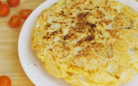 Tortilla vegana dorada y esponjosa, con trozos de patata visibles, servida en plato blanco. Tomates cherry al lado.