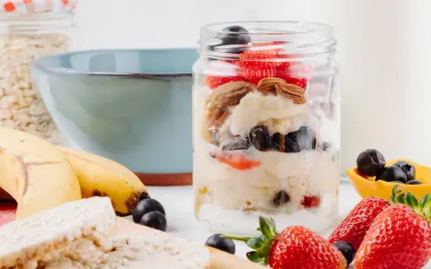 Deliciosa avena nocturna en un frasco con fresas, arándanos y almendras. Plátanos y galletas de arroz al lado.