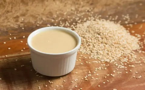 Un cuenco blanco con cremosa salsa de tahini junto a una pila de semillas de sésamo sobre una tabla de madera.