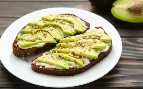 Dos tostadas de pan integral con queso crema, rodajas de aguacate y semillas. Un desayuno saludable y delicioso.