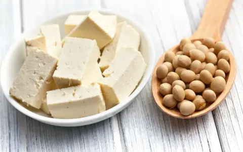 Tofu en cubos en un plato blanco junto a una cuchara de madera llena de semillas de soja. ¿A que sabe el tofu? Neutro, absorbe sabores.