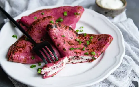 Crepes rosados ​​rellenos de queso crema, perfectos para recetas con remolacha cruda. Decorados con cebollín y acompañados de una porción de crema agria.