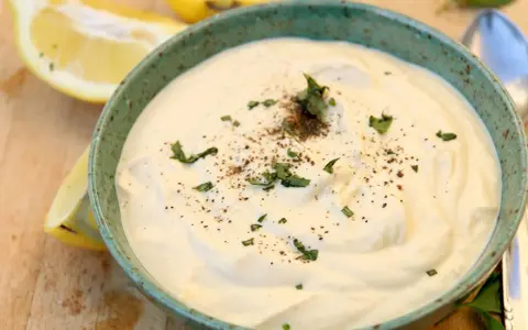 Un cremoso tazón de mayonesa de tofu, adornado con perejil fresco y pimienta negra, junto a rodajas de limón.