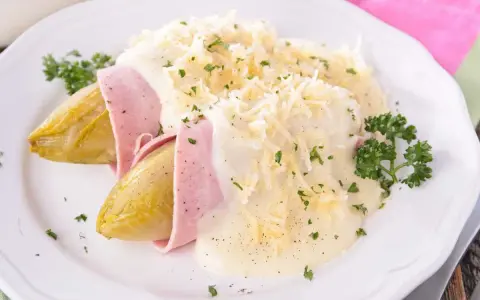 Endivias al horno envueltas en jamón, cubiertas con una cremosa salsa bechamel y queso rallado, adornadas con perejil fresco.
