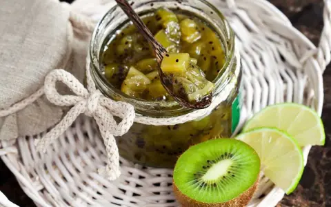 Tarro de mermelada de kiwi casera, con una cuchara de madera llena de la dulce conserva. Rodajas de kiwi y lima acompañan la presentación.