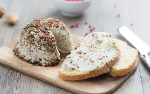 Queso vegano receta: un bloque de queso cremoso cubierto de pimienta y hierbas, servido con rebanadas de pan.