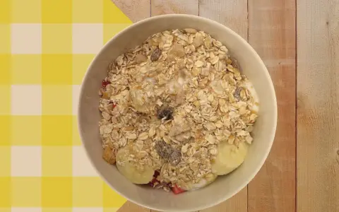 Un tazón de porridge de avena con plátano, fresas y pasas, servido sobre un mantel a cuadros amarillos y blancos.