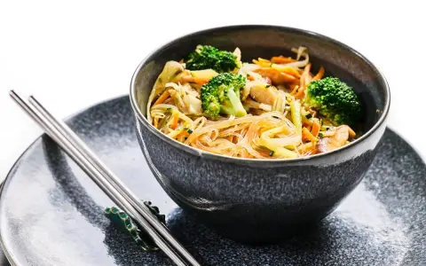Deliciosa receta con noodles de arroz, brócoli, zanahoria y otros vegetales frescos en un tazón gris, acompañado de palillos.