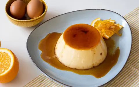 Delicioso flan de naranja con caramelo líquido, decorado con rodajas y ralladura de naranja. Huevos en un cuenco al fondo.