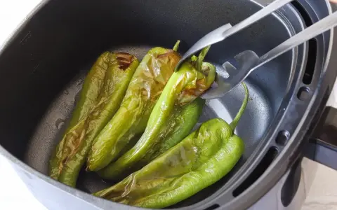 Pimientos verdes asándose en una freidora de aire. Unas pinzas metálicas sujetan uno de los pimientos, mostrando su piel ligeramente chamuscada.