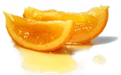 Dos rodajas de naranja confitada brillan con dulzura, bañadas en un almíbar dorado sobre fondo blanco.