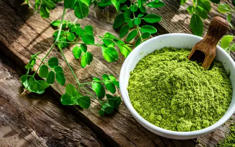 Polvo verde vibrante de planta moringa en un cuenco blanco, junto a ramas frescas de la planta moringa sobre madera rústica.