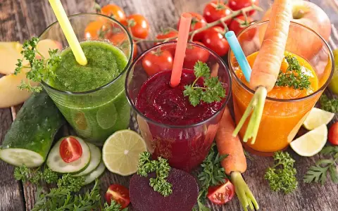 Tres zumos astringentes de colores vibrantes: verde, rojo y naranja, rodeados de ingredientes frescos como zanahorias, remolachas y tomates.